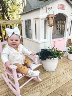 a baby sitting in a pink rocking chair on a wooden deck next to a doll house