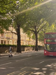 a red double decker bus driving down a street next to tall trees and people on scooters