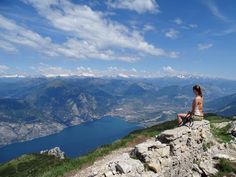 a woman sitting on top of a rock cliff looking at the mountains and lake below