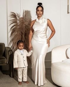 a woman standing next to a little boy in a white dress and suit with one hand on his hip