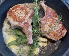 two pieces of meat are cooking in a skillet with vegetables and herbs on the side