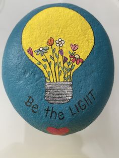 a painted rock that says be the light with flowers in a vase on it and words below