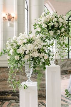 two tall vases with white flowers and greenery are on pedestals in front of a mirror