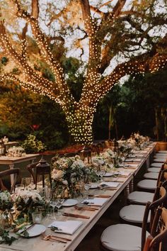 an outdoor dinner table set up with place settings and lights on the trees in the background