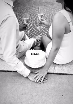 a man and woman sitting on the ground with a cake in front of them, holding wine glasses