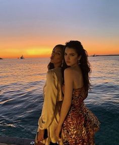 two young women standing next to each other near the ocean at sunset or sunrise with boats in the background