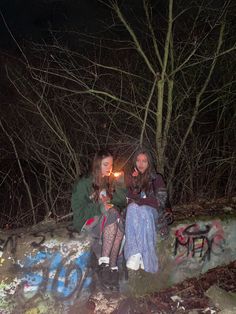 two women sitting on a rock with graffiti all over it and trees in the background