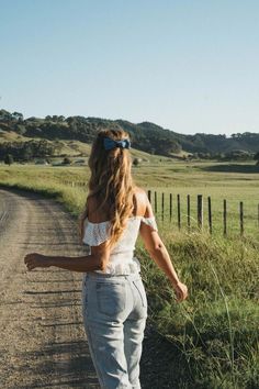 a woman walking down a dirt road next to a lush green field