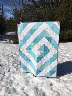 a blue and white quilt hanging from a clothes line in the snow with trees in the background