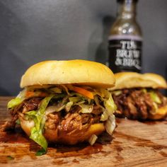 two pulled pork sandwiches sitting on top of a wooden cutting board next to a bottle of beer