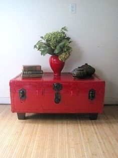 a red trunk sitting on top of a hard wood floor next to a vase filled with flowers