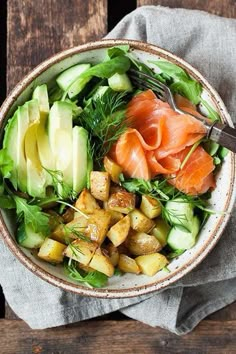 a bowl filled with vegetables and salmon on top of a wooden table next to a fork