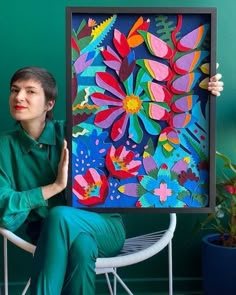 a woman sitting in a chair holding up a large colorful painting on the wall above her head