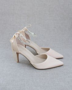 a pair of wedding shoes with bows on the heel and beaded ankle straps, sitting against a gray background