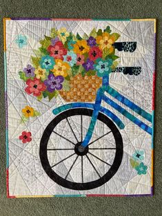 a quilted wall hanging with a bicycle and flowers in the basket on it's front wheel