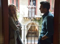 a man and woman standing in front of a balcony looking at each other with their arms crossed