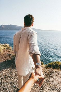 a man and woman holding hands while standing on top of a hill next to the ocean