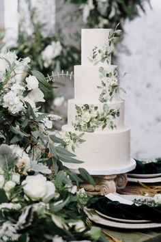 a wedding cake with greenery and white flowers on the table next to plates, forks and napkins