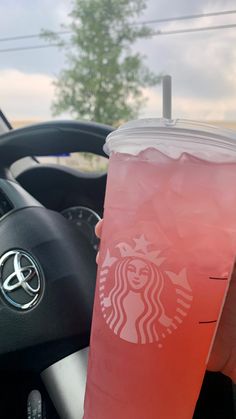 a person holding up a drink in front of a car steering wheel with the driver's hand