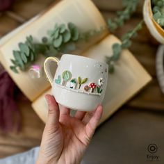 A hand holds a whimsical white mug featuring a 3D forest scene with trees, mushrooms, and a small house. Open books and greenery are in the background. Whimsical Mushrooms, Coffee Experience, Enchanted Garden, Botanic Garden, Gold Handles, Tea Or Coffee, Lush Garden, Nature Lovers, Handmade Ceramic