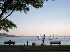 people sitting on benches near the water with sailboats in the background and one person walking