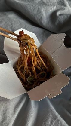 an open box with noodles and chopsticks in it on top of a bed