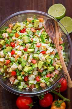 a glass bowl filled with chopped vegetables and avocado next to sliced tomatoes, lime wedges and two wooden spoons