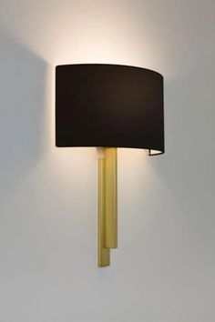 A modern wall sconce with a black drum shade and gold vertical mount, emits a soft, warm glow against a white wall, creating an elegant ambiance.