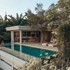 a woman sitting on the edge of a swimming pool next to a house with trees