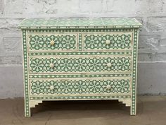 a green and white painted dresser against a brick wall with an intricate design on the drawers