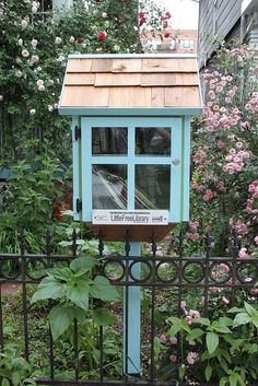 a blue birdhouse sitting on top of a metal fence next to flowers and trees