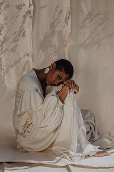 a woman sitting on top of a bed covered in white sheets with her hands under her chin