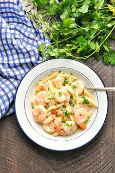 a white bowl filled with pasta and shrimp on top of a wooden table next to herbs