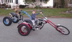 two children are sitting on small motorcycles in the parking lot with one child standing next to them