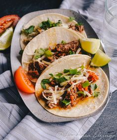 three tacos on a plate with limes and tomatoes