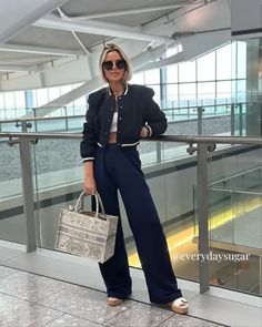 a woman standing on a balcony holding a handbag and wearing sunglasses, with her hands in her pockets