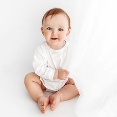 a smiling baby sitting on the floor wearing a white bodysuit and looking at the camera