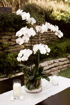 white flowers are in a glass vase on a table with candles and rocks behind it