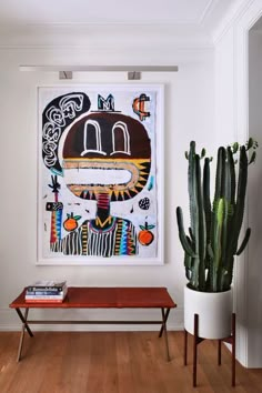 a painting hangs on the wall next to a wooden bench and cactus in a white room