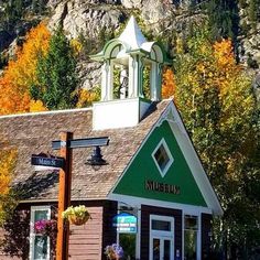 a small building with a clock tower on the top in front of mountains and trees