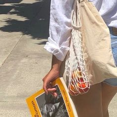 a woman is walking down the street with a book in her hand and a bag full of oranges