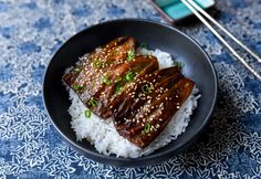 a black plate topped with rice and meat next to chopsticks on a blue table cloth