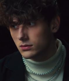 a young man with curly hair wearing a white turtle neck sweater and pearls on his necklace