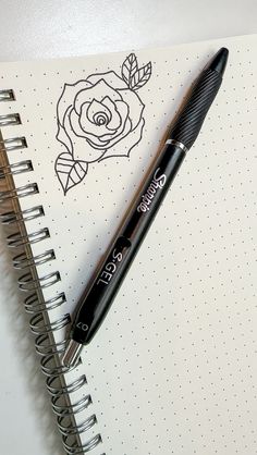 a black pen sitting on top of an open spiral notebook next to a drawing of a rose