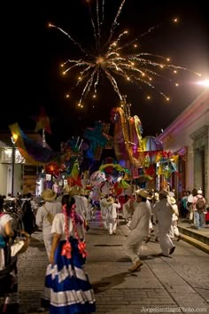 many people are walking down the street at night with fireworks in the sky above them