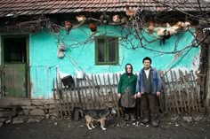 a man and woman standing in front of a blue house with chickens hanging on the wall