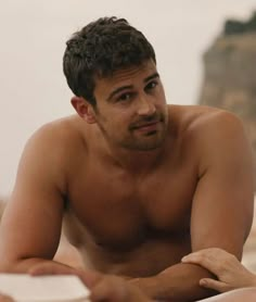 a shirtless man sitting on the beach looking at something in his hand while reading a book