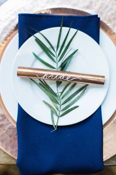 a place setting with blue napkins, gold chargers and greenery on the plate