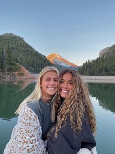 two women hugging each other in front of a lake with mountains and trees behind them