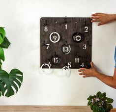 a man holding up a wooden clock with numbers on it and two hands next to a potted plant